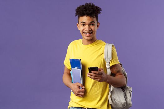 Back to school, university concept. Portrait of young handsome smiling man, student asking for classmate phone number, making note in mobile phone, wear backpack, hold notebooks and study material