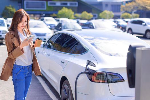 Smiling young woman standing on city parking near electric car, charging automobile battery from small city station, drinking coffee and using smartphone