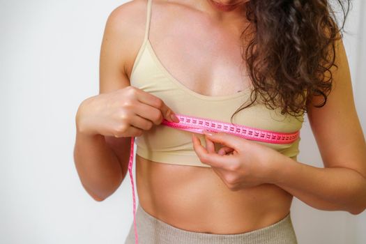 Cropped view of slim woman measuring breasts with tape measure at home, close up. Unrecognizable European woman checks the result of a weight loss diet or liposuction indoors. Healthy lifestyle.