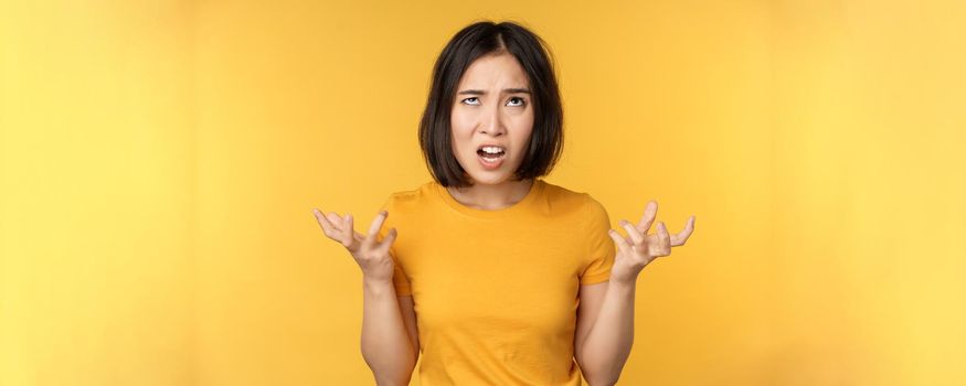 Image of angry asian woman, shouting and cursing, looking outraged, furious face expression, standing over yellow background