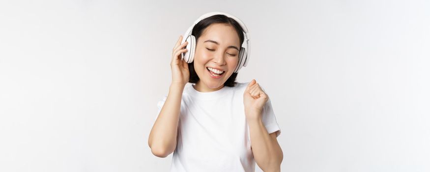 Modern asian girl dancing, listening music with headphones, smiling happy, standing in tshirt over white background
