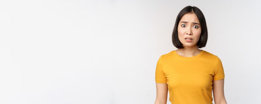 Portrait of worried korean girl, looking concerned at camera, standing in yellow tshirt over white background