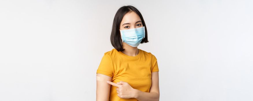 Covid-19, vaccination and healthcare concept. Portrait of cute asian girl in medical mask, has band aid on shoulder after coronavirus vaccine, standing over white background