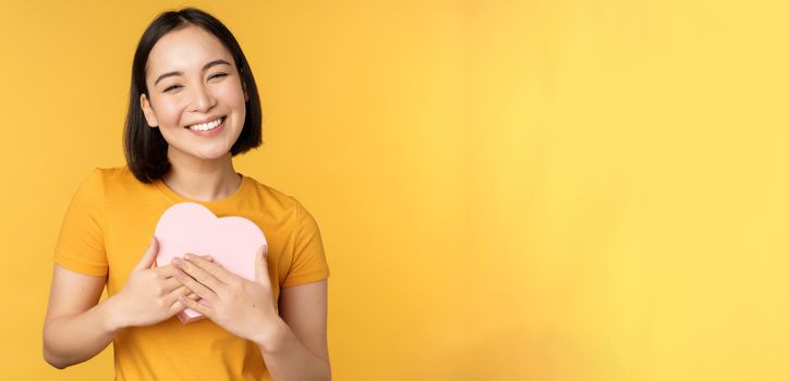 Romance and valentines day. Happy beautiful asian woman holding big heart card and smiling, standing over yellow background.
