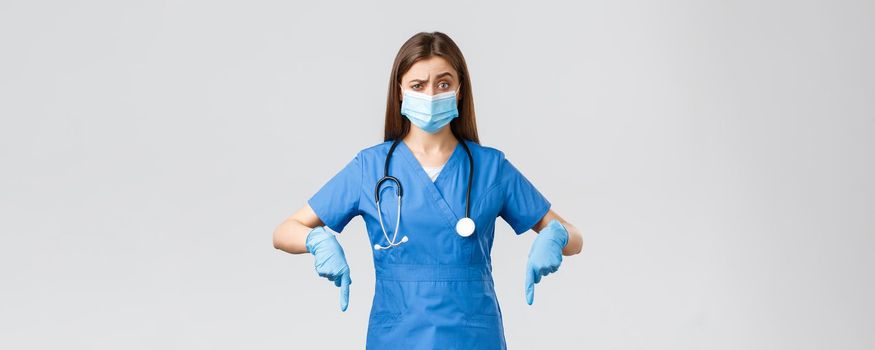 Covid-19, preventing virus, health, healthcare workers and quarantine concept. Skeptical and unsure female nurse or doctor in blue scrubs, personal protective equipment, pointing fingers down unsure