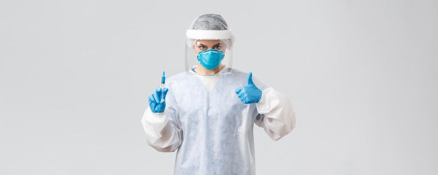 Covid-19, medical research, healthcare workers and quarantine concept. Confident professional doctor in personal protective equipment PPE costume, show thumb-up, hold syringe with corona vaccine