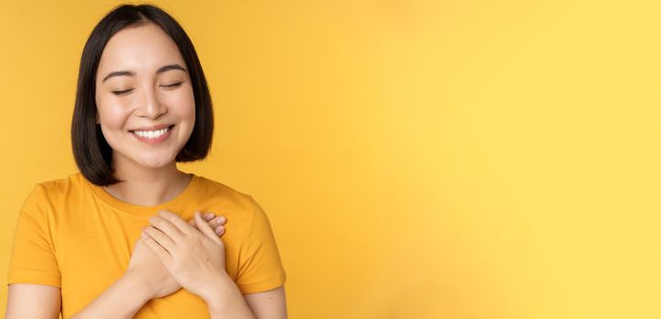 Beautiful asian woman, smiling with tenderness and care, holding hands on heart, standing in tshirt over yellow background