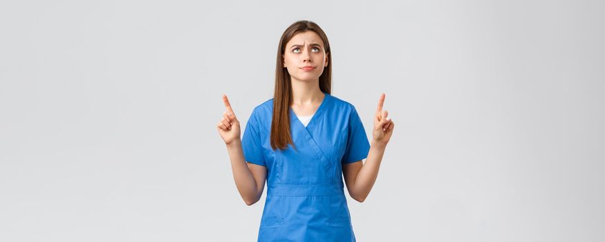 Healthcare workers, prevent virus, covid-19 test screening, medicine concept. Dissatisfied and upset young moody nurse, doctor in blue scrubs, smirk and frowning, pointing finger up