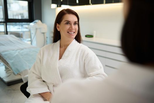 Attractive middle-aged Caucasian woman in white terry bathrobe during a visit to wellness spa clinic for receiving beauty anti-aging treatment on her face and body. Cosmetology and medical concept