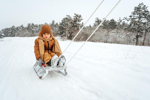 Little toddler child in winter outfit sits on sledge in snowy park