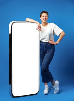 Happy young woman leaning on huge cellphone with empty white screen, for your ad, mock up