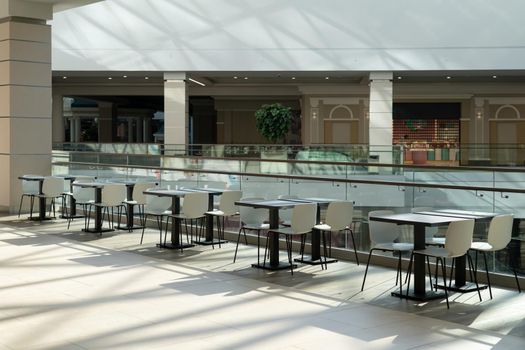 A row of tables with white chairs for visitors to the food court of a modern shopping center.