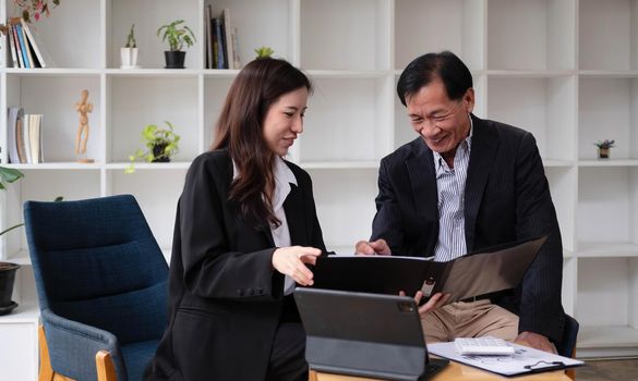 Happy woman and middle-aged man sit at office desk work on laptop together laughing on funny joke, smiling diverse colleagues have fun talking cooperating at workplace