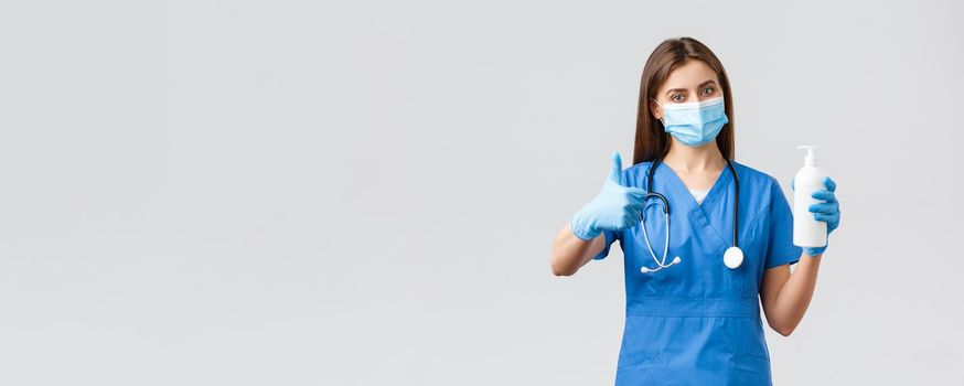 Covid-19, preventing virus, health, healthcare workers and quarantine concept. Confident female doctor, nurse in blue scrubs in medical mask and scrubs advice using hand sanitizer, thumbs-up approval