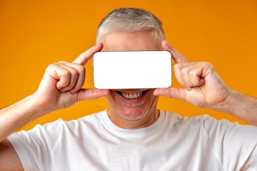 Middle age man holding smartphone on face over yellow background