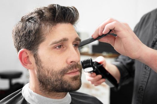Man barber cutting hair of male client with clipper at barber shop. Hairstyling process.