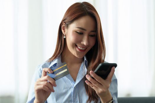 Young beautiful Asian woman using smartphone and credit card for online shopping at home with copy space. E-payment technology, shopaholic lifestyle, or mobile phone financial application concept