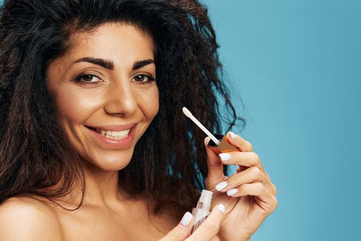 Close up portrait of cheerful happy tanned pretty curly Latin female hold lipstick smile at camera posing isolated over pastel blue background. Cosmetic lips product ad Natural beauty Makeup concept