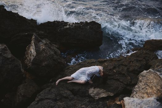 Barefoot woman in long white dress wet hair lying on a rocky cliff view from above