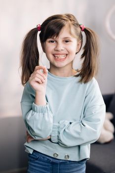 Portrait of cute little child girl is rejoices and showing her lost milk tooth and smiling to camera of toothless mouth while she standing in bedroom. Happy girl holding her fallen tooth in hand.