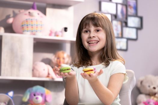 Attractive smiling cute school kid girl holds four tasty cookies in her hands and look at camera. Traditional French multicolored macaroon. Food concept