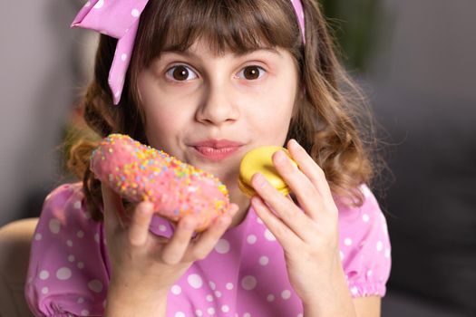 Adolescent school girl plays with sweet donuts doing happy fun face expressions on background. Cute little girl in pink dress holding macaron and donut in hands by the face. Funny concept with sweets