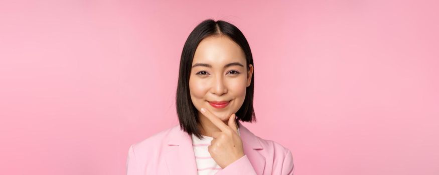 Close up portrait of smiling asian working lady in suit, businesswoman looking thoughtful, thinking or deciding smth, standing over pink background