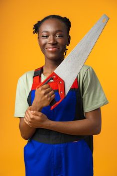 Portrait of a cute african american woman wearing uniform posing with a wood saw in her hands against yellow background
