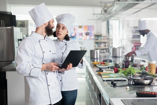 Food industry workers standing in restaurant professional kitchen while using computer to decide dinner service dish.
