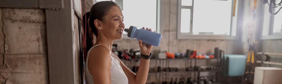 Slim woman in sportswear in the gym holding flask of water, smiling