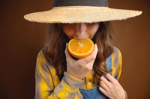 Studio portrait of an attractive caucasian young woman in a hat and denim overalls holds a cut orange fruit in her hands and shows her tongue trying to taste it
