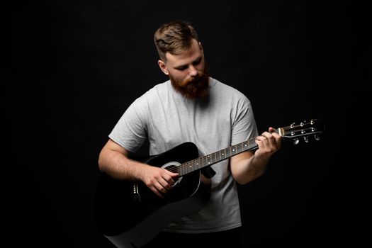 Handsome brunette bearded guitarist plays an acoustic guitar in a black room. The concept of music recording, rehearsal or live performance.