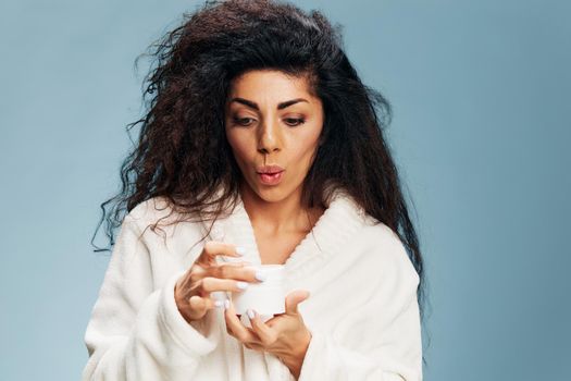 Excited shocked happy curly Latin lady in bathrobe open beauty cream in white jar, closing eyes, posing isolated on pastel blue background. Cosmetic product ad concept. Good offer. Copy space