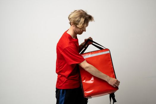 Young courier opening thermal bag on white background. Food delivery service. Delivery guy in a red t-shirt uniform work as courier and holds red thermal food backpack. Service concept.