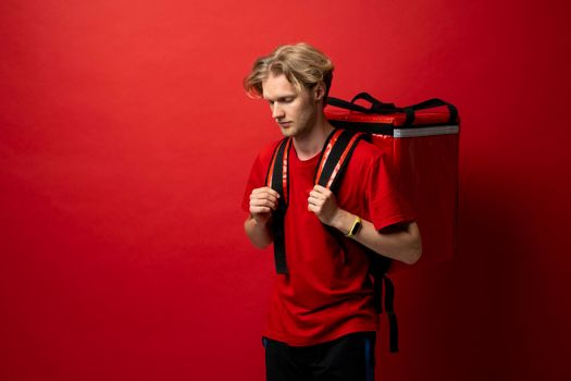 Young delivery employee man in red tshirt uniform with a thermal food bag backpack works as a courier in a food delivery service on a white background.