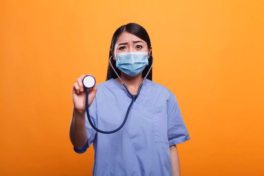 Worried concerned healthcare clinic asian nurse wearing protective facemask while holding stethoscope. Distressed caretaker wearing virus protection mask while holding medical instrument.