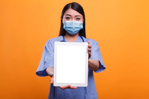 Healthcare clinic asian nurse wearing virus protection facemask and stethoscope while holding a tablet in front of camera. Caregiver holding touchscreen device while wearing blue uniform and mask.