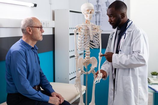 Physiotherapist doctor showing anatomical human skeleton explaining back pain to old man patient