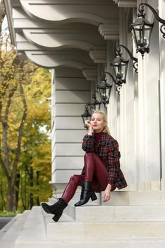 A beautiful, modern, fashionable blonde girl with a red lipstick posing outdoors . red leather leggings, turtleneck and checkered jacket. fit figure.