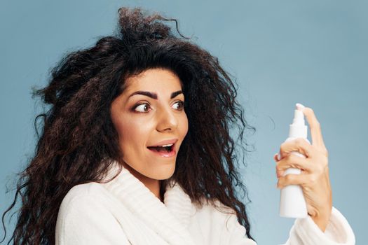 Shocked smiling curly Latin lady in bathrobe look at toner lotion spray bottle in hand, open mouth, posing isolated on pastel blue background, excited of effective skincare routine. Copy space