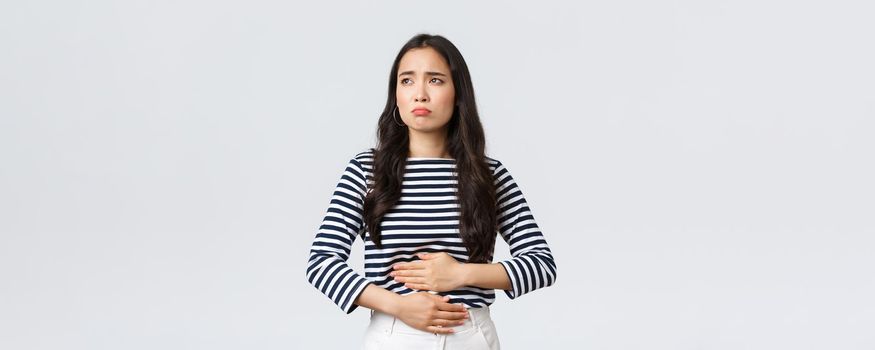 Lifestyle, beauty and fashion, people emotions concept. Woman got food poisoned, touching belly feeling unwell. Asian girl with cramps looking gloomy, having menstrual pain, white background
