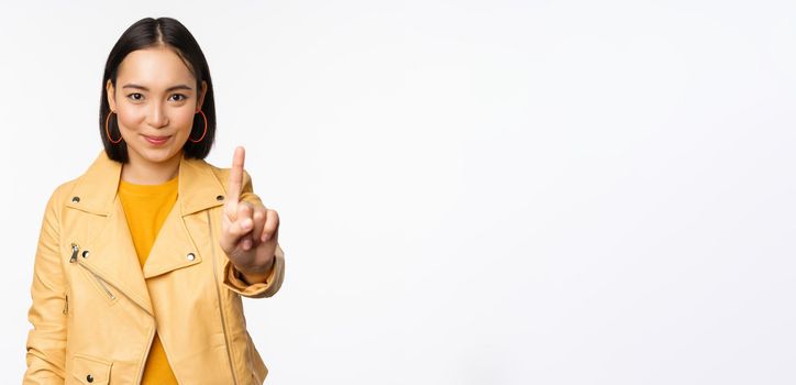 Rule number one. Confident smiling asian woman showing index finger, stop, prohibit gesture, disapprove smth, standing over white background