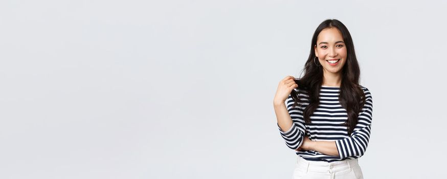Lifestyle, people emotions and casual concept. Charming smiling korean girl in striped shirt, touching hair strand and grinning happily camera, talking to friend, white background