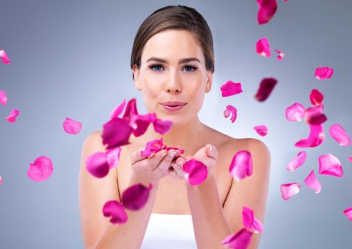 Blossom into a new you. An attractive young woman blowing rose petals against a grey background.