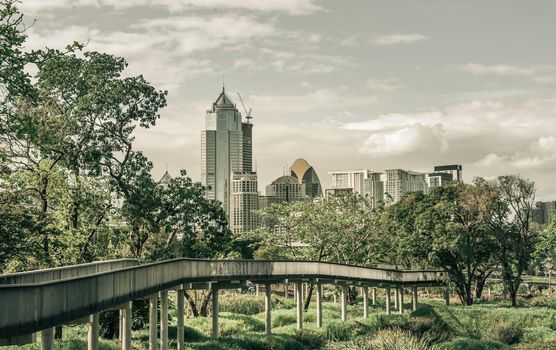 Bangkok, Thailand - Feb 19, 2022 : View of Modern high buildings and walkway bridge among surrounded green trees space against blue sky with clouds at afternoon. City growth concept, Selective focus.