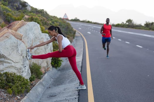 Fit african american woman in sportswear stretching while man is running on a coastal road