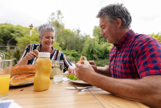 Smiling caucasian senior man holding hamburger eating meal with wife in garden