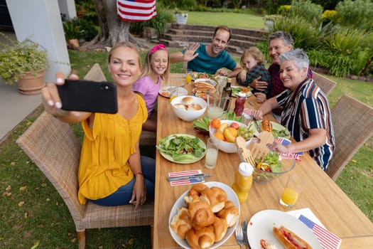 Caucasian woman taking selfie with three generation family having celebration meal in garden. three generation family celebrating independence day eating outdoors together.