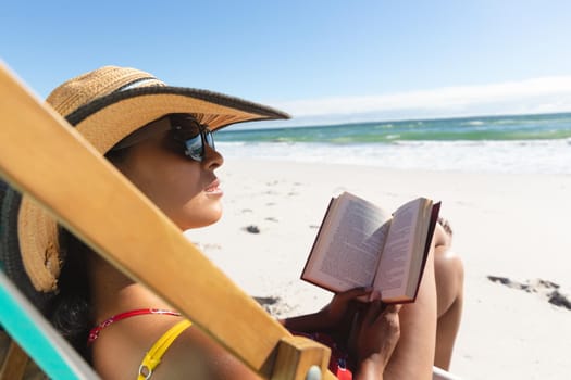 Mixed race woman on beach holiday sitting in deckchair reading book
