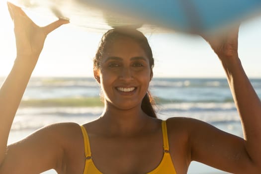 Happy mixed race woman on the beach carrying surfboard on head